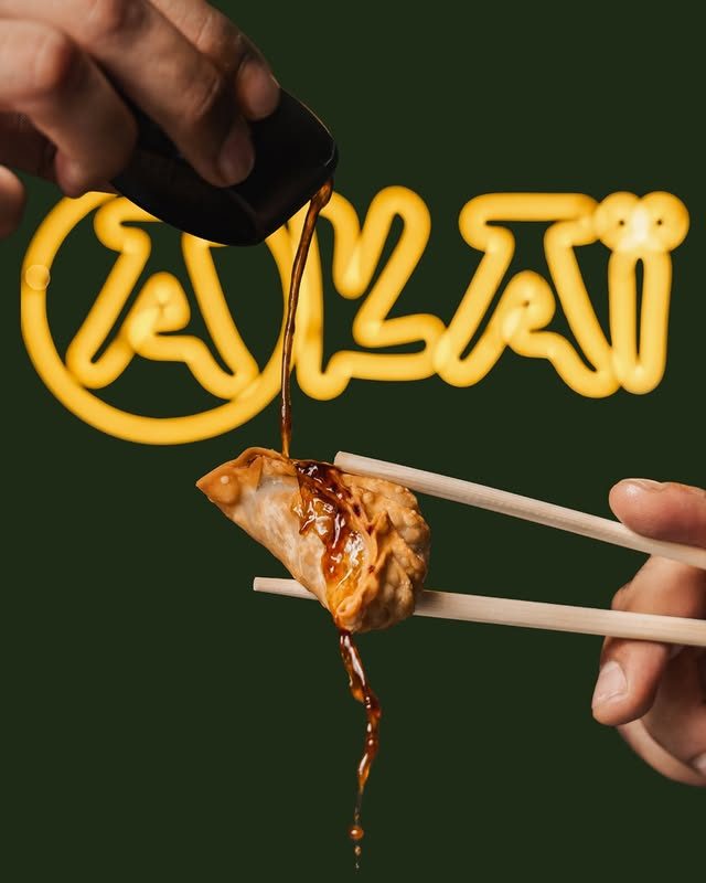 Gyoza croustillant saisi aux baguettes, nappé de sauce soja devant l'enseigne néon AKAI